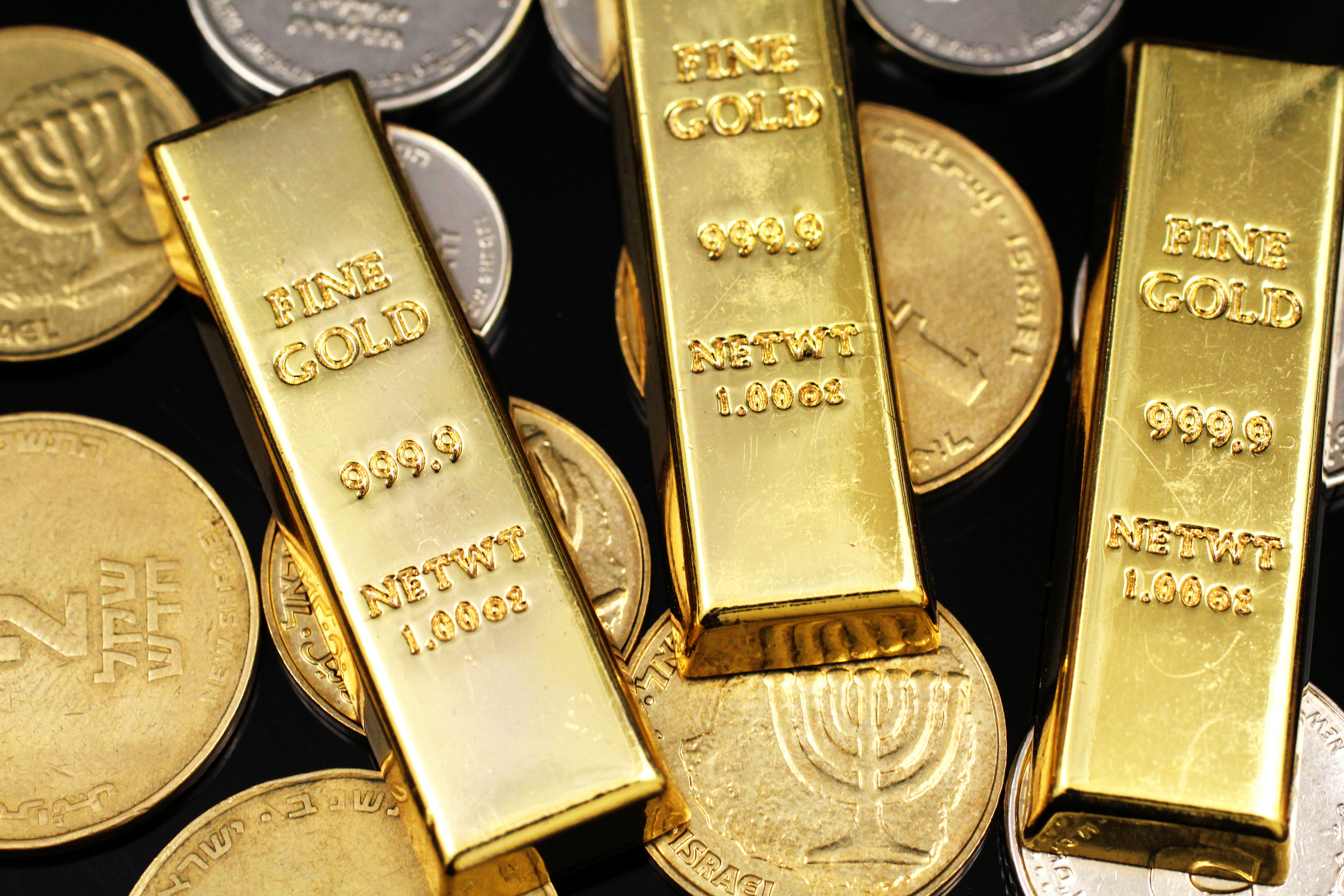 A close up image of an assortment of Israeli coins with three small gold ingots on a black background